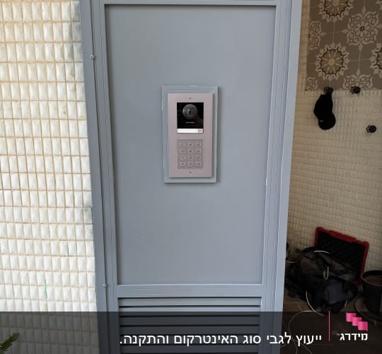 אינטרקום עם מצלמה ולוח מקשים על דלת מתכת אפורה